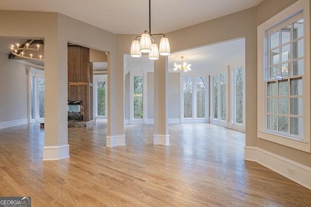 unfurnished dining area featuring ornate columns, a multi sided fireplace, light wood-type flooring, and an inviting chandelier