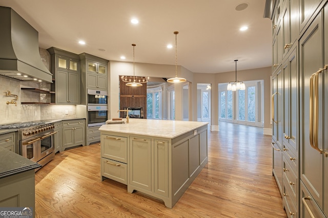 kitchen featuring premium range hood, appliances with stainless steel finishes, decorative light fixtures, dark stone countertops, and a kitchen island with sink