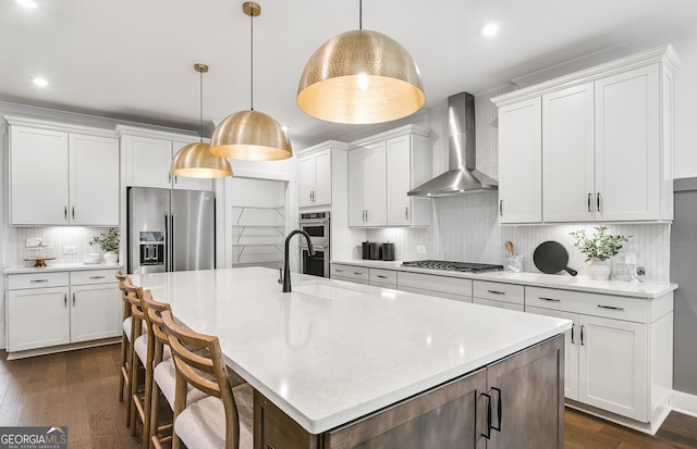 kitchen with white cabinetry, a center island with sink, appliances with stainless steel finishes, pendant lighting, and wall chimney range hood