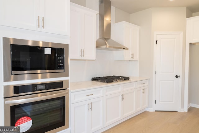 kitchen with light wood finished floors, light countertops, appliances with stainless steel finishes, white cabinetry, and wall chimney exhaust hood