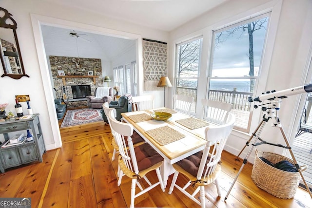 dining area featuring a stone fireplace, a water view, wood-type flooring, vaulted ceiling, and ceiling fan