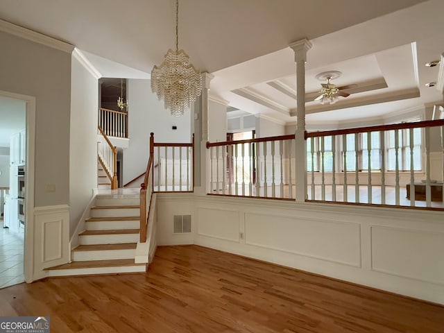 staircase featuring crown molding, hardwood / wood-style flooring, ceiling fan with notable chandelier, a raised ceiling, and ornate columns