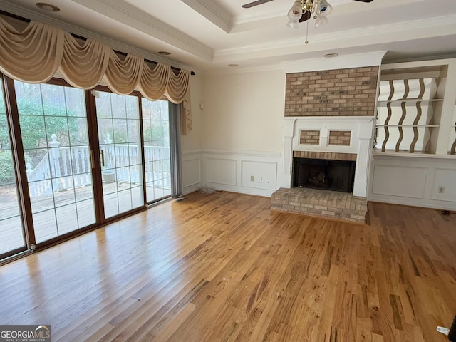 unfurnished living room with a brick fireplace, ornamental molding, a raised ceiling, ceiling fan, and light hardwood / wood-style floors