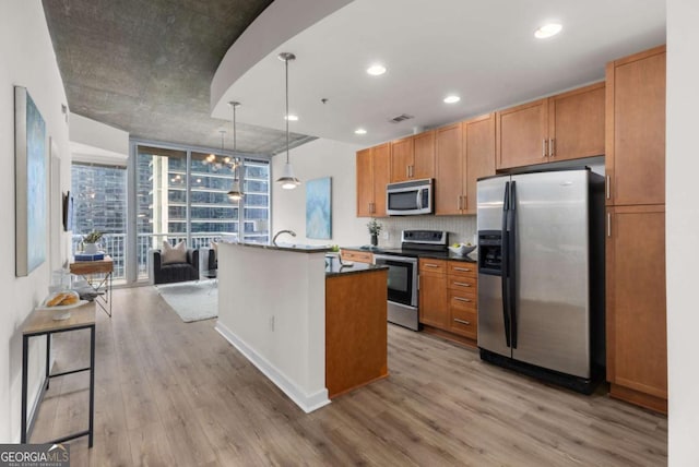 kitchen with appliances with stainless steel finishes, decorative light fixtures, decorative backsplash, a center island with sink, and light wood-type flooring