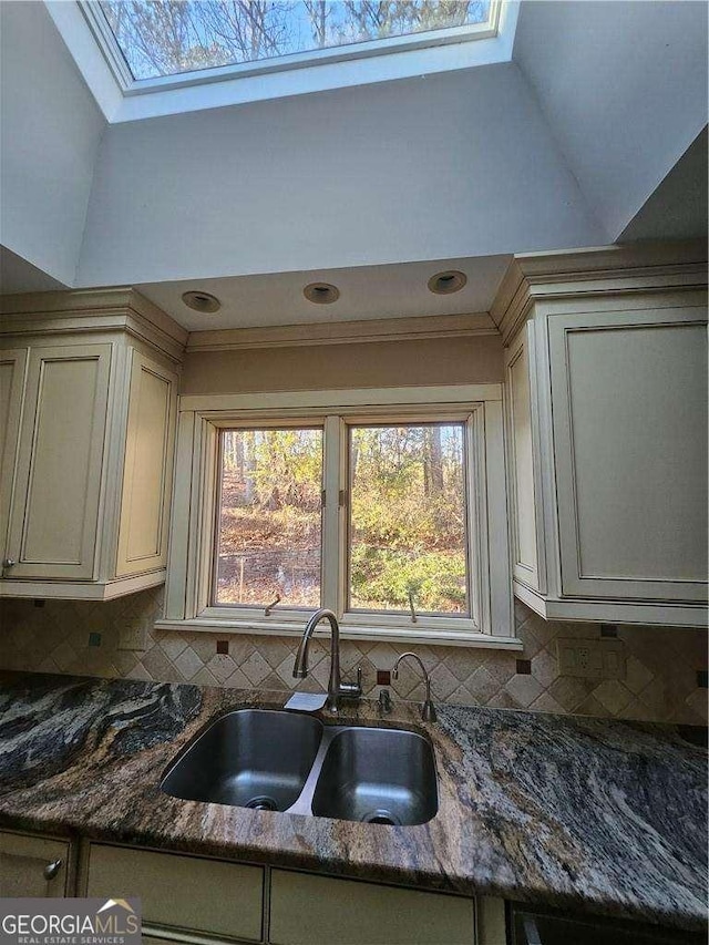 kitchen featuring vaulted ceiling with skylight, sink, dark stone countertops, decorative backsplash, and cream cabinetry