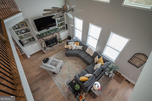 living room with a brick fireplace, built in features, hardwood / wood-style flooring, ceiling fan, and a high ceiling