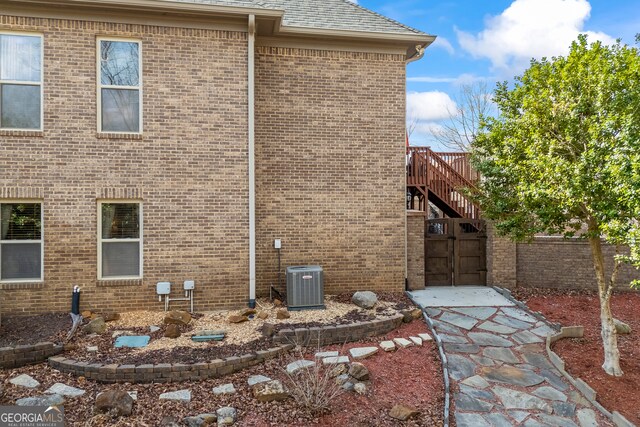 view of home's exterior with central AC, brick siding, a shingled roof, fence, and stairway