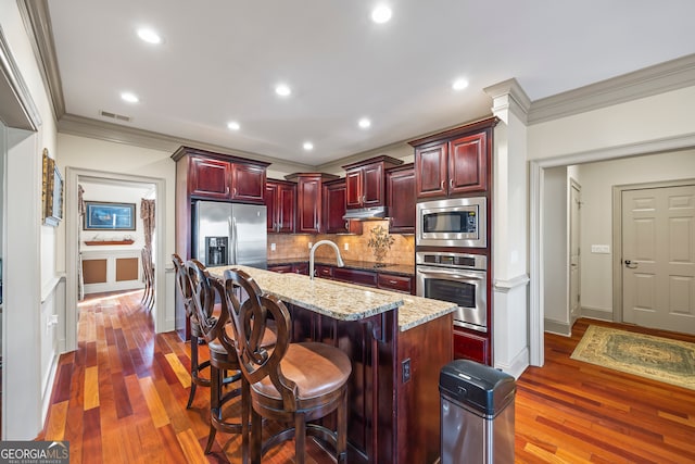 kitchen with backsplash, stainless steel appliances, light stone countertops, an island with sink, and a kitchen bar