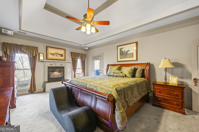 bedroom with ornamental molding, a tray ceiling, a glass covered fireplace, and light colored carpet