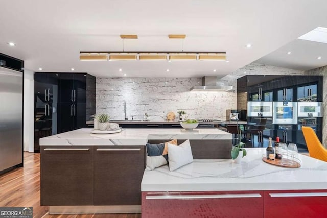 kitchen featuring a spacious island, decorative light fixtures, stainless steel refrigerator, and wall chimney exhaust hood