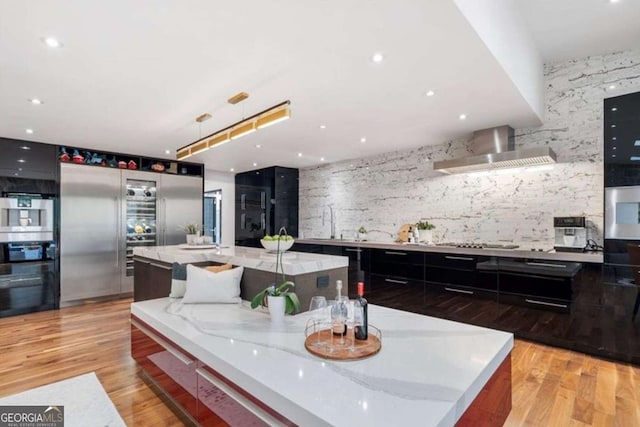 kitchen with a spacious island, wall chimney exhaust hood, light hardwood / wood-style floors, and light stone counters