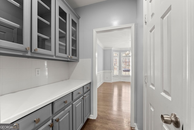 interior space with dark wood-style floors, a wainscoted wall, gray cabinetry, glass insert cabinets, and ornamental molding