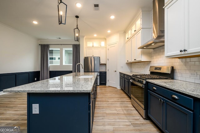 kitchen with hanging light fixtures, stainless steel appliances, an island with sink, white cabinets, and wall chimney exhaust hood