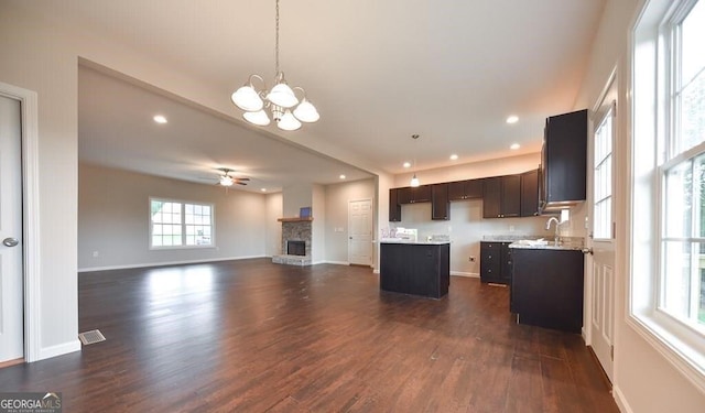 kitchen with a stone fireplace, a center island, dark hardwood / wood-style floors, pendant lighting, and ceiling fan with notable chandelier