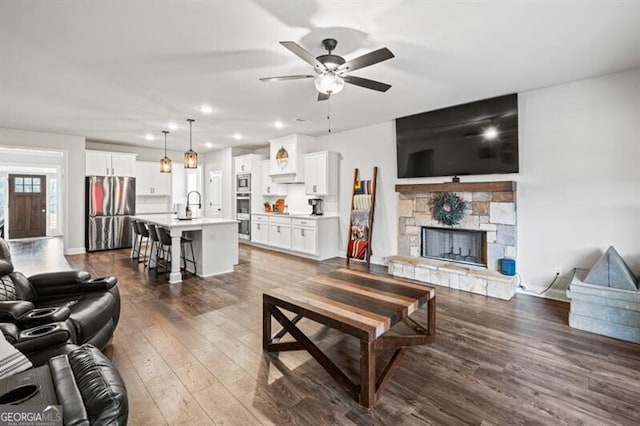 living room featuring a stone fireplace, dark hardwood / wood-style floors, sink, and ceiling fan