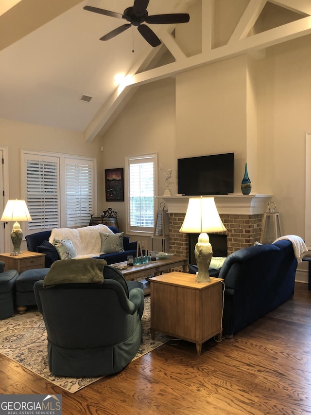 living room with ceiling fan, high vaulted ceiling, wood-type flooring, a brick fireplace, and beamed ceiling