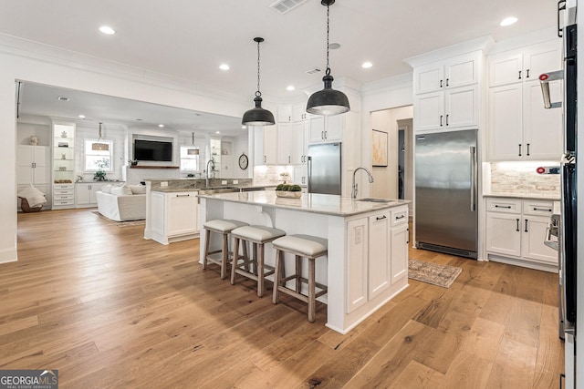 kitchen featuring stainless steel built in refrigerator, decorative light fixtures, an island with sink, and white cabinets