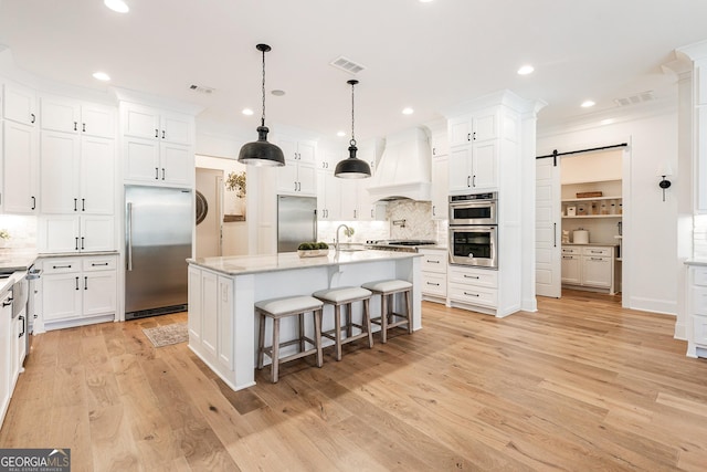 kitchen featuring appliances with stainless steel finishes, white cabinetry, premium range hood, an island with sink, and a barn door