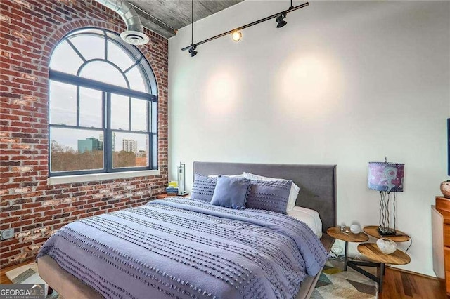 bedroom featuring rail lighting, wood finished floors, and brick wall