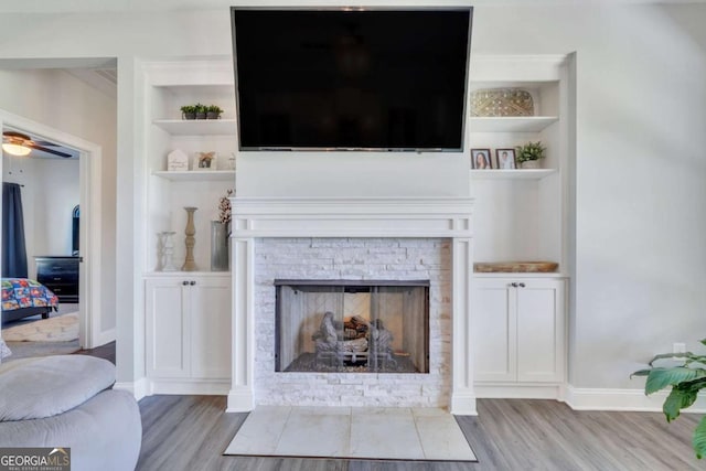 living room featuring built in shelves, ceiling fan, a stone fireplace, and light wood-type flooring