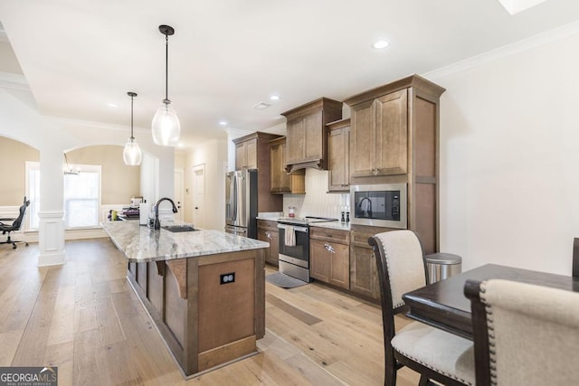 kitchen featuring arched walkways, an island with sink, stainless steel appliances, crown molding, and a sink