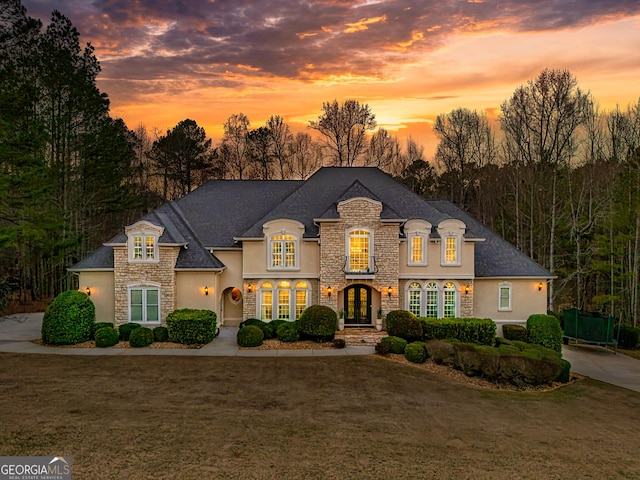 french country inspired facade featuring stucco siding, stone siding, french doors, and a yard