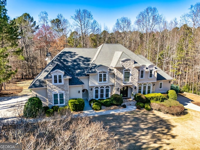 french provincial home featuring a forest view, stone siding, a chimney, and stucco siding