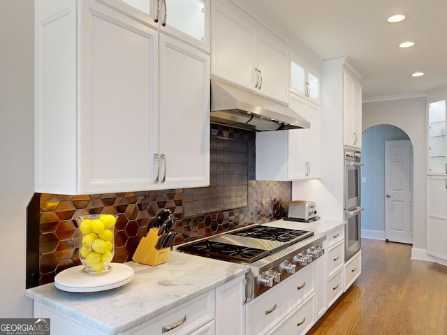 kitchen with arched walkways, under cabinet range hood, stainless steel appliances, white cabinetry, and glass insert cabinets