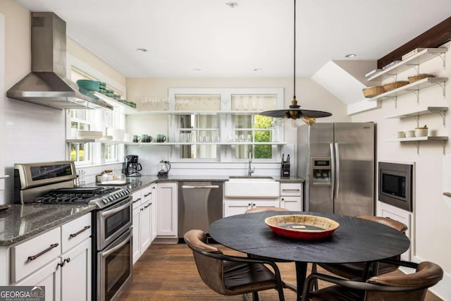 kitchen with dark stone counters, appliances with stainless steel finishes, wall chimney range hood, open shelves, and a sink