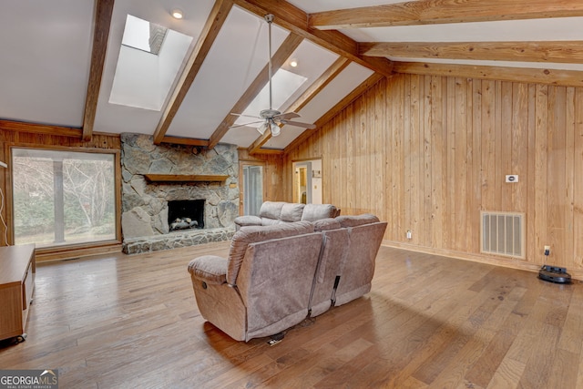 living room featuring a stone fireplace, wooden walls, a skylight, beamed ceiling, and light hardwood / wood-style floors