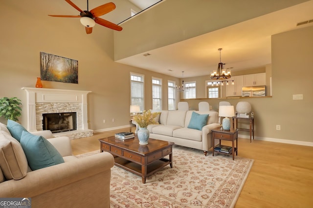 living room featuring ceiling fan with notable chandelier, a stone fireplace, light hardwood / wood-style floors, and a high ceiling