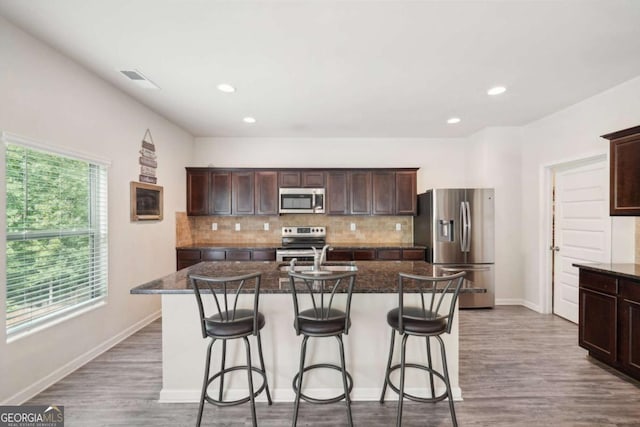 kitchen with dark stone countertops, appliances with stainless steel finishes, an island with sink, hardwood / wood-style flooring, and decorative backsplash