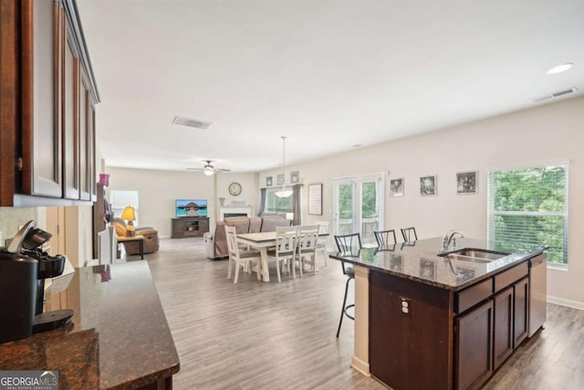 kitchen with sink, dark brown cabinets, a center island with sink, light wood-type flooring, and dark stone counters