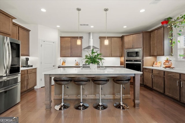 kitchen with wall chimney range hood, stainless steel appliances, hanging light fixtures, and a center island with sink