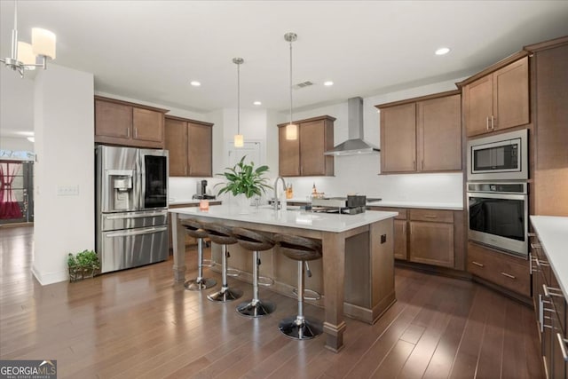 kitchen with appliances with stainless steel finishes, a kitchen bar, hanging light fixtures, a center island with sink, and wall chimney range hood