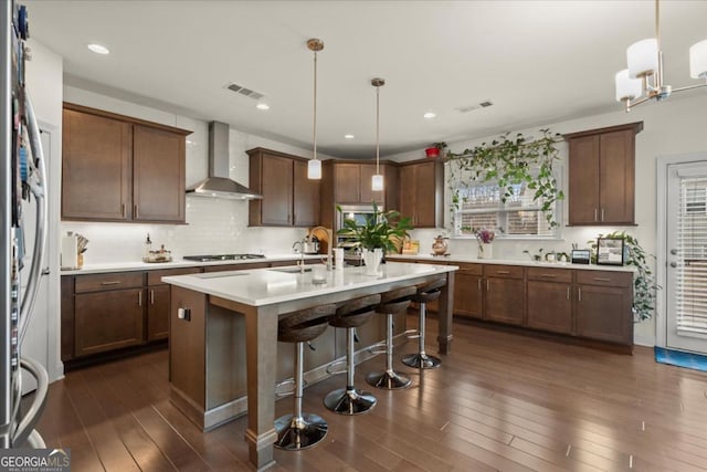 kitchen with hanging light fixtures, an island with sink, wall chimney exhaust hood, and appliances with stainless steel finishes