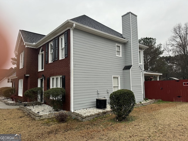 view of side of property with fence, central AC unit, a shingled roof, and a chimney