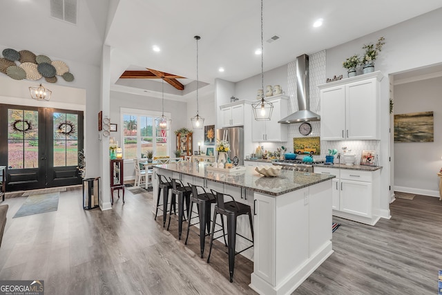 kitchen featuring wall chimney exhaust hood, white cabinetry, a center island with sink, stainless steel refrigerator, and dark stone counters