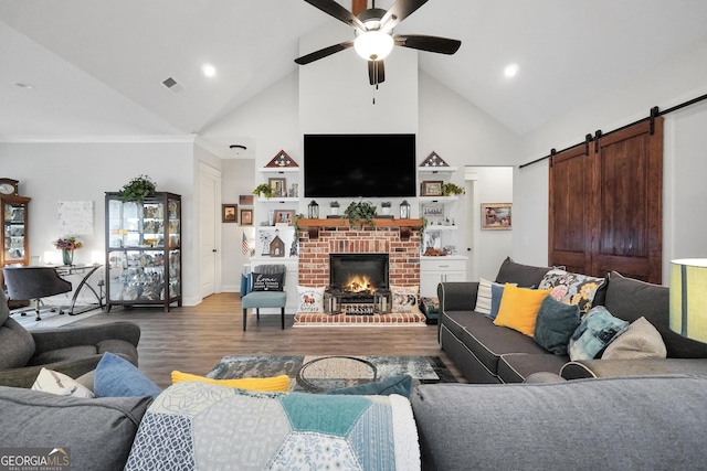 living room with ceiling fan, hardwood / wood-style floors, high vaulted ceiling, a brick fireplace, and a barn door