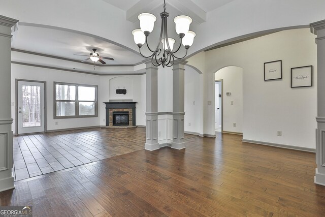 unfurnished living room featuring decorative columns, arched walkways, a fireplace with raised hearth, dark wood-type flooring, and ceiling fan with notable chandelier