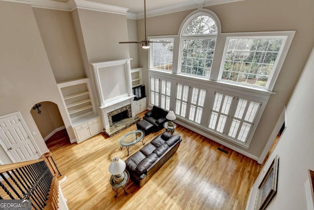 living room with built in features, a high ceiling, wood-type flooring, ornamental molding, and a stone fireplace