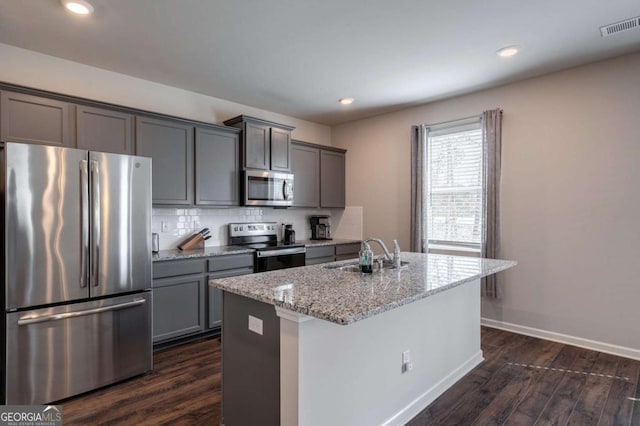 kitchen with appliances with stainless steel finishes, dark hardwood / wood-style floors, sink, a kitchen island with sink, and light stone countertops