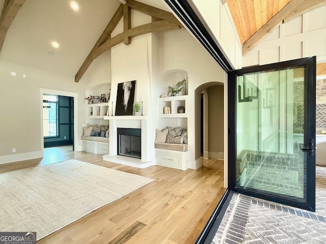 interior space featuring baseboards, arched walkways, beamed ceiling, light wood-type flooring, and built in shelves