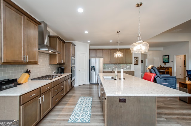 kitchen featuring appliances with stainless steel finishes, an island with sink, hanging light fixtures, light stone counters, and wall chimney exhaust hood