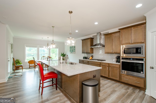 kitchen with appliances with stainless steel finishes, decorative light fixtures, an island with sink, ornamental molding, and wall chimney exhaust hood