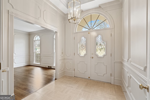 foyer entrance featuring visible vents, a decorative wall, an inviting chandelier, ornamental molding, and light wood-style floors