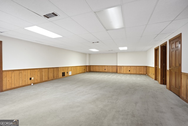 empty room with light carpet, wooden walls, visible vents, a drop ceiling, and wainscoting