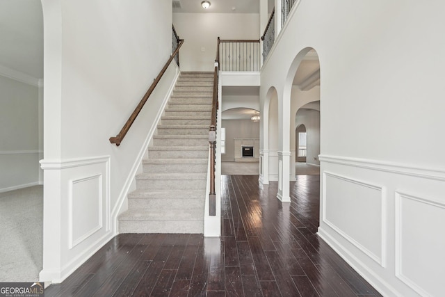 staircase featuring a fireplace with raised hearth, a decorative wall, a high ceiling, wainscoting, and wood finished floors