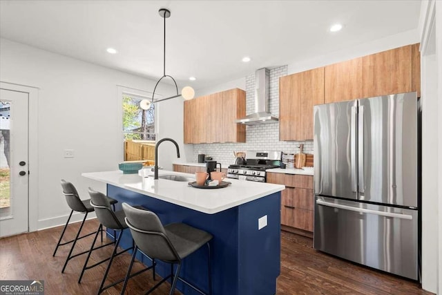 kitchen featuring an island with sink, a sink, light countertops, appliances with stainless steel finishes, and wall chimney range hood