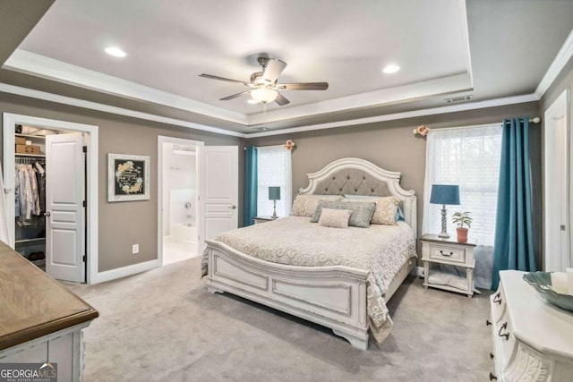 carpeted bedroom featuring connected bathroom, ornamental molding, a walk in closet, ceiling fan, and a tray ceiling